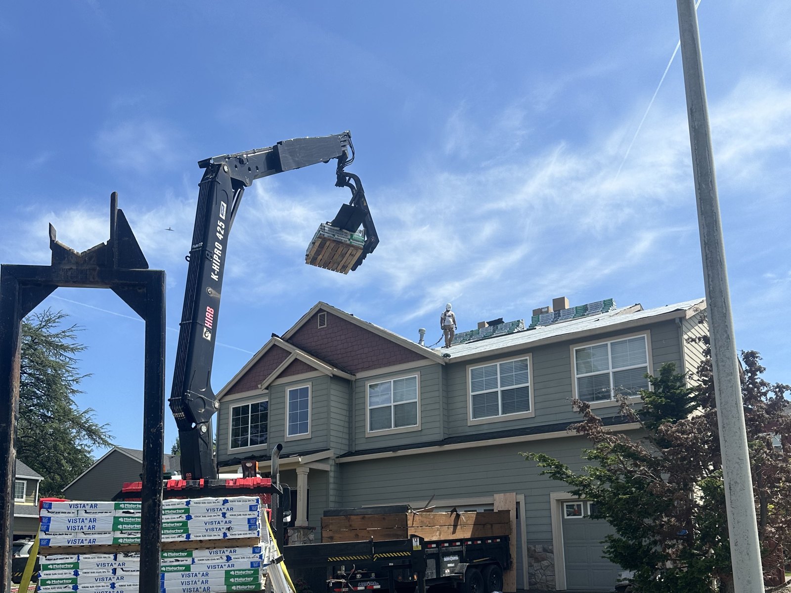 Boom truck lifting a bundle of shingles onto a roof mid-replacement while a roofer works on the stripped decking